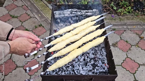 Close-up of hands turning skewers with sulguni cheese wrapped in golden puff pastry dough, baking on charcoal grill with smoke and flame