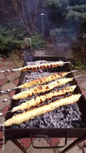 Close-up of hands turning skewers with sulguni cheese wrapped in golden puff pastry dough, baking on charcoal grill with smoke and flame