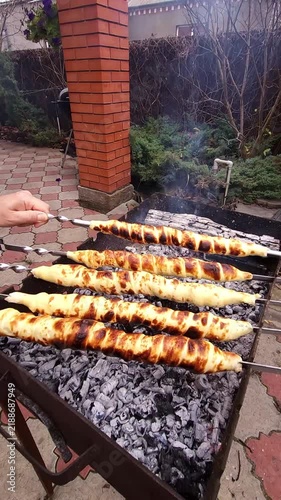 Close-up of hands turning skewers with sulguni cheese wrapped in golden puff pastry dough, baking on charcoal grill with smoke and flame