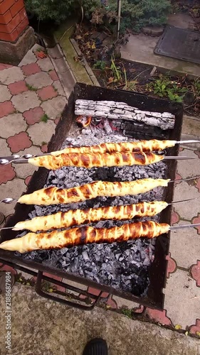 Close-up of hands turning skewers with sulguni cheese wrapped in golden puff pastry dough, baking on charcoal grill with smoke and flame