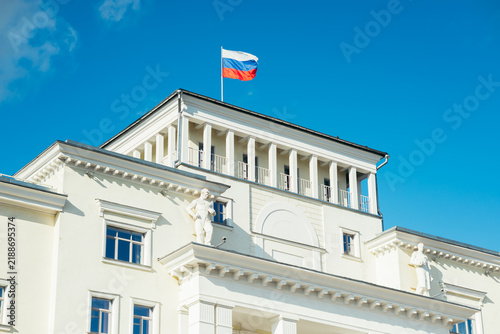 A Russian flag is displayed on the roof of a white government building. The building features ornate architectural details and stands against a bright blue sky