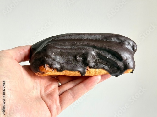 Quadro su tela A person holds a delicious chocolate-glazed eclair in their hand against a neutral white background
