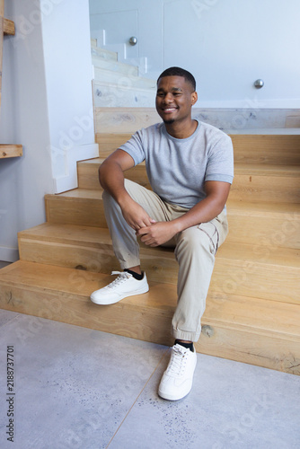 African American adult man sitting on light-wood staircase at home, smiling, wearing white sneakers