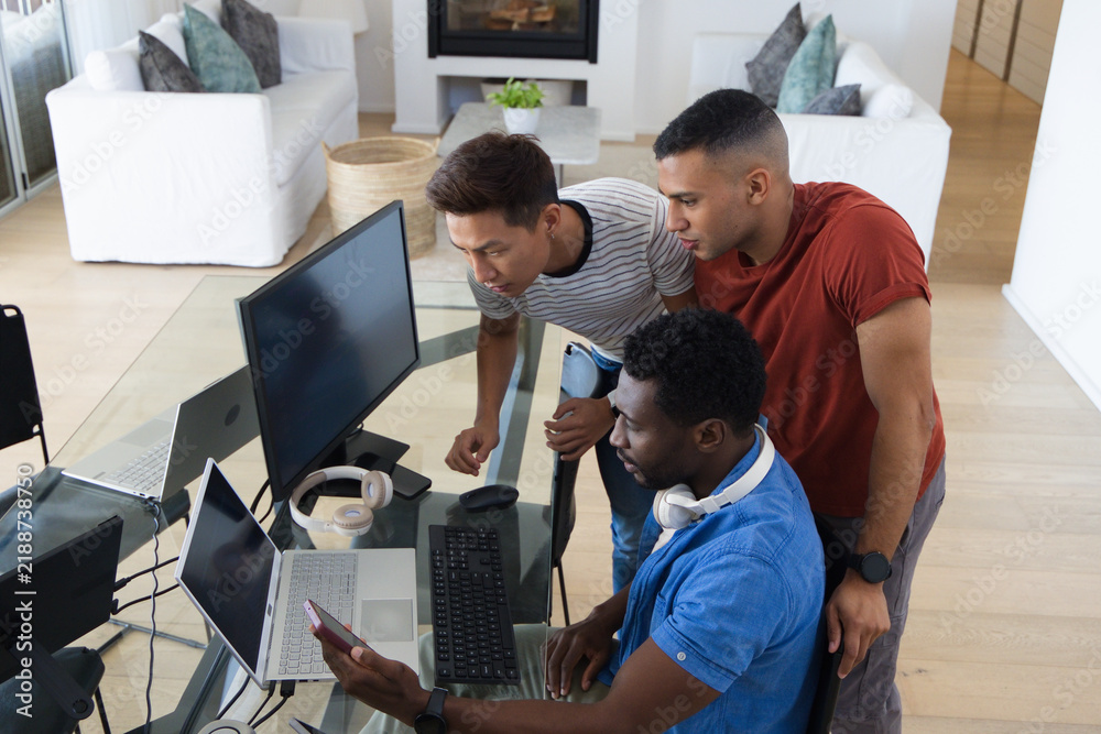 Fototapeta premium Diverse male coworkers leaning over glass desk in living room workspace, viewing phone and laptops