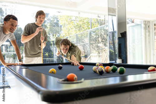 Diverse adult male friends taking shot over pool table with billiard balls by glass doors