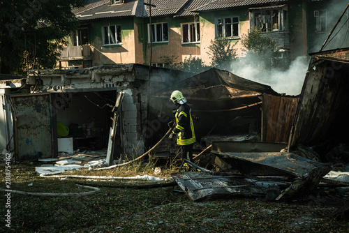 Konstfotografi Ukrainian firefighter holding a hose line next to smoldering and destroyed garage boxes with a damaged apartment building in the background