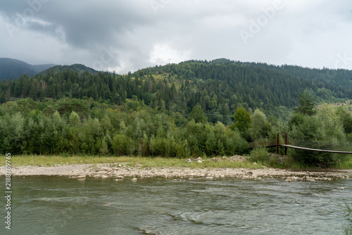 Scenic wild mountain river with a suspension footbridge in the Carpathian Mountains, Ukraine. Authentic rural nature landscape, eco-tourism destination, and misty alpine pine forest under a stormy sky
