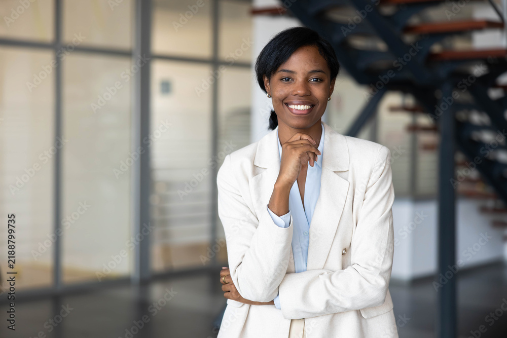 Obraz premium Black business woman standing confidently in a modern office environment, smiling at camera with hand on chin