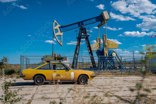 Vintage yellow coupe with a rooftop cargo box parked beside an old oil pumpjack behind a fence, under a bright blue sky with scattered clouds. Rustic industrial roadside scene.