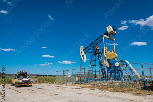 Vintage yellow coupe with a rooftop cargo box parked beside an old oil pumpjack behind a fence, under a bright blue sky with scattered clouds. Rustic industrial roadside scene.