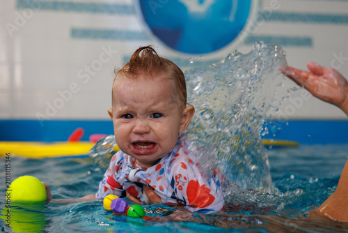 Photography Water splashes behind a upset infant holding colorful toys during swimming class