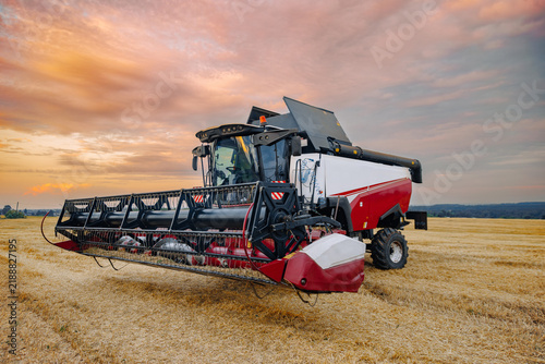 Agricultural combine harvester parked in a golden wheat field under a colorful sunset sky with rolling hills visible in the background