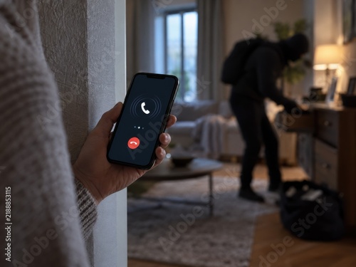 A person hiding behind a wall watches a masked burglar rummaging through items while on a phone.