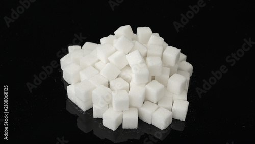 Konstfotografi Pile of white sugar cubes on a glossy black surface, with faint reflections and scattered sugar granules around the base