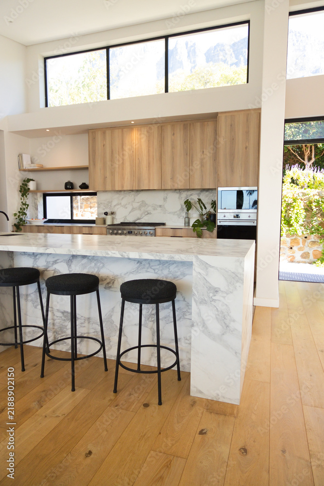 Obraz premium Marble-look waterfall kitchen island featuring three black stools and glass door opening to garden