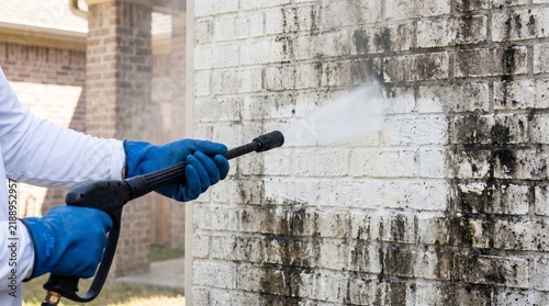 Pressure washer operator hands, with water spray cleaning mold grime, washing brick wall, residential home exterior, home maintenance photography