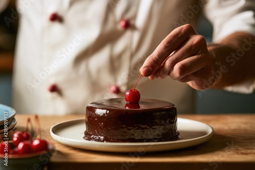 Chef's hands place a cherry atop a glossy chocolate cake, a sweet finish