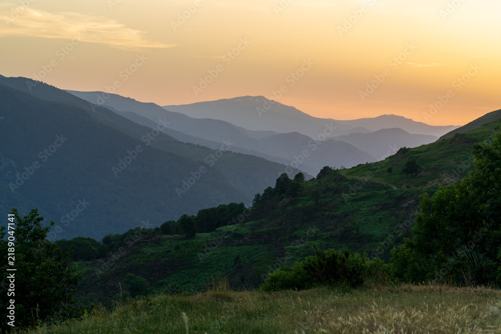 Obraz premium Rolling ridgelines stretch toward a glowing orange horizon as dusk settles over a scenic alpine mountain valley. Dense green shrubs and trees cover the foreground hills beneath the fading daylight