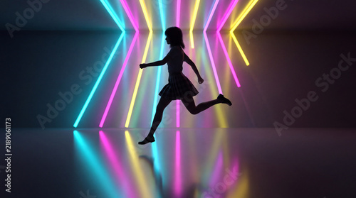A silhouette of a girl in a skirt running past bright blue, yellow, and pink neon light tubes in a dark, reflective studio environment.