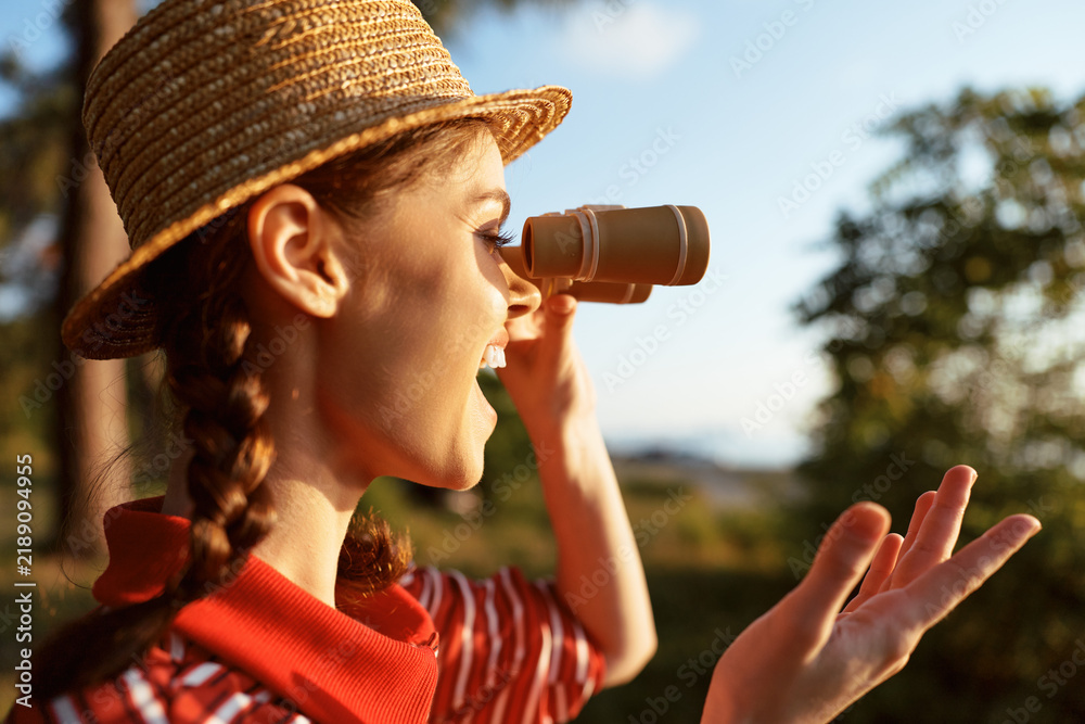 Obraz premium A happy young woman in a straw hat holding binoculars exploring nature lifestyle. She enjoys sunny weather and outdoor activity during a summer trip for travel and adventure.