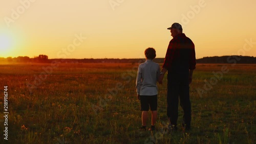 Grandfather and child standing together in open field at golden sunset, sharing peaceful moment while watching horizon and warm evening light in countryside
