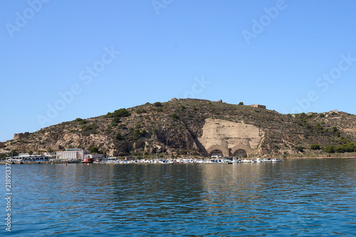 A sunny day view of a cliffside harbor with docked boats, coastal architecture, and a clear blue sky in Cartagena, Murcia, Spain.