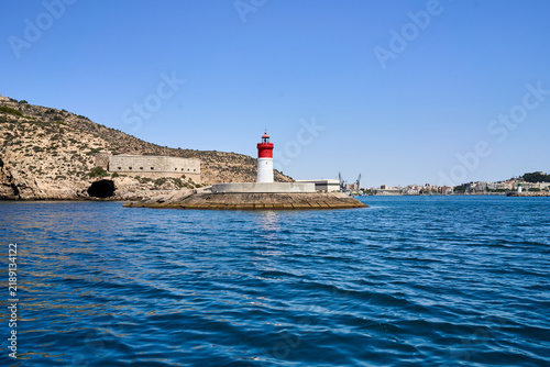 The iconic lighthouse stands guard near the coast, near a big mountain and some constructions, under a clear blue sky on a sunny day.