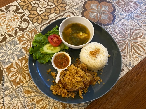 Konstfotografi Indonesian fried chicken meal served with steamed rice, soup, chili paste, cucumber, and lettuce on a dark plate on a wooden table