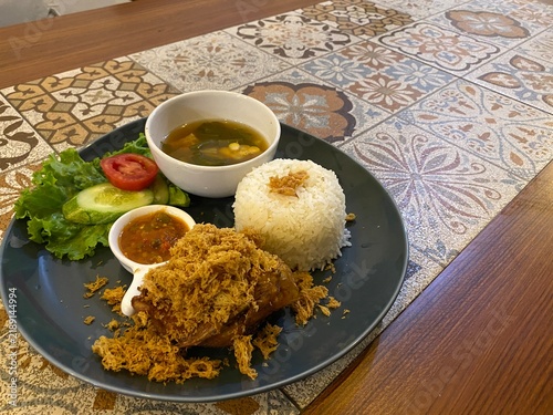 Canvastavla Indonesian fried chicken meal served with steamed rice, soup, chili paste, cucumber, and lettuce on a dark plate on a wooden table