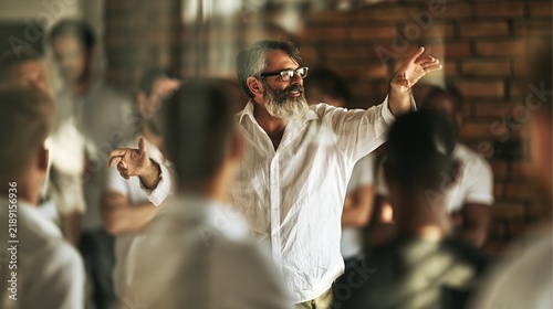 A bearded man passionately speaks to an engaged audience in a bright, modern setting.