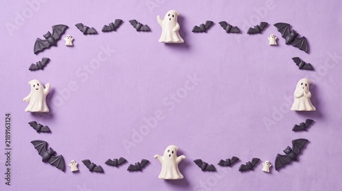 A festive halloween frame made of small white ghost figurines and black bat decorations on a solid purple background surface