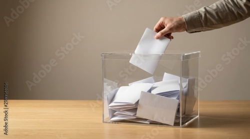 Person putting a paper ballot into a clear acrylic voting box on a wooden table