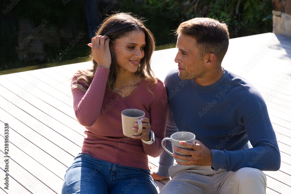 Obraz premium Diverse couple sitting on wooden deck by pond, holding ceramic mugs and wearing smartwatch