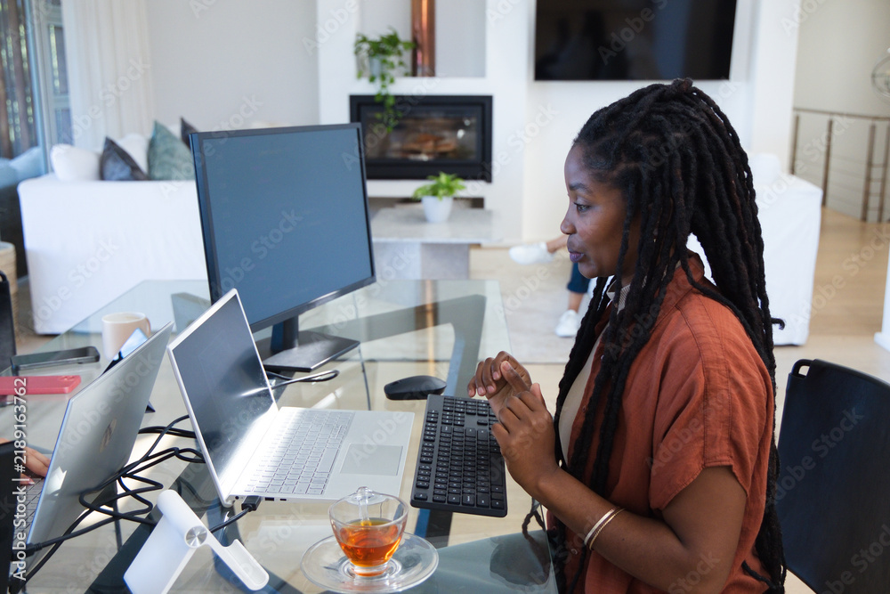 Obraz premium African American woman sitting at glass-top desk in living room using laptop and monitor