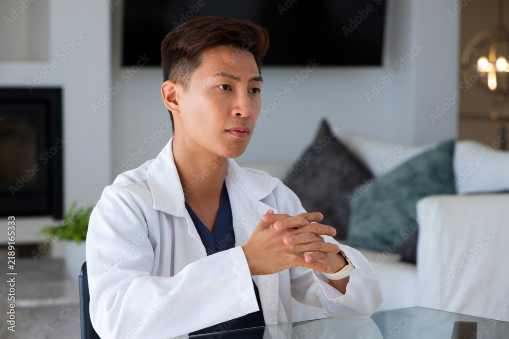 Obraz premium Mid-adult Asian man sitting at glass-topped table at home wearing white lab coat over navy scrubs