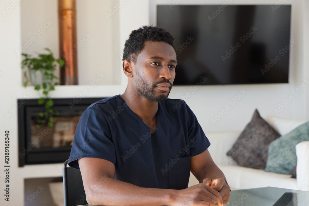 Obraz premium Mid-adult African man sitting at glass table in living room wearing navy scrubs, viewing TV