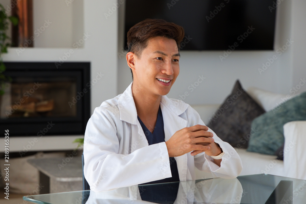 Obraz premium Mid adult Asian man sitting at glass-topped table in modern consultation room, wearing lab coat