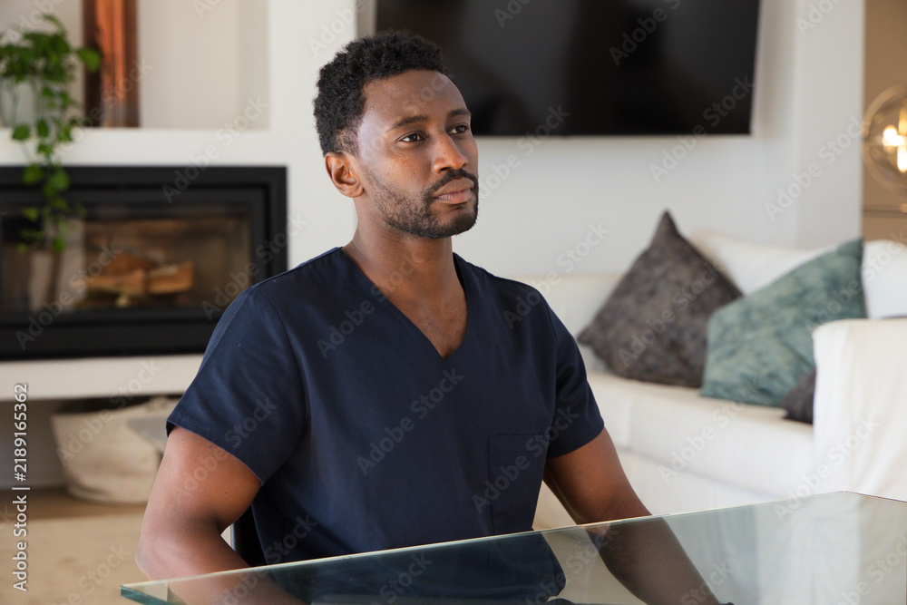 Obraz premium Mid adult African man sitting at glass-topped table in modern living room wearing navy scrub top
