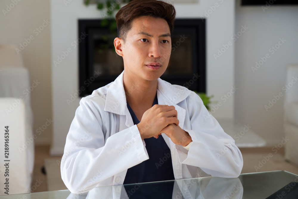 Obraz premium Asian man sitting in home office wearing white lab coat and dark scrubs at glass table