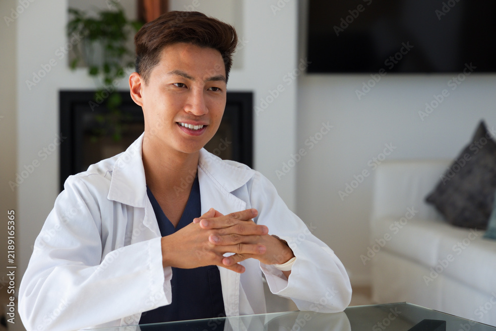 Obraz premium Mid-adult Asian man wearing white lab coat, navy scrubs sitting at glass table in living room
