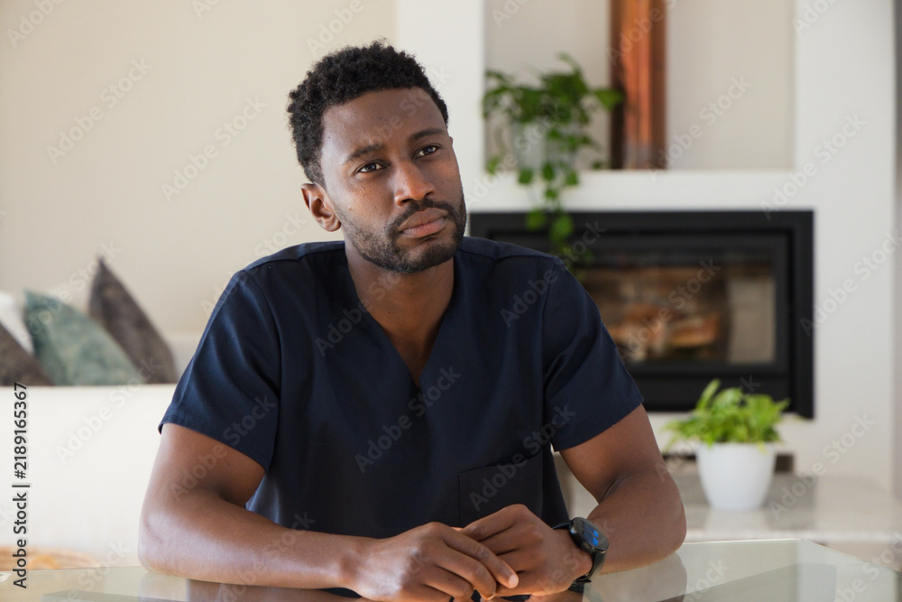 Fototapeta premium Mid adult African man wearing navy scrub top, sitting at table in living room, clasping hands