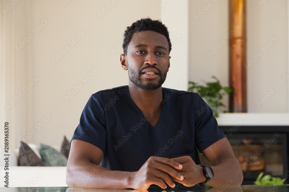 Obraz premium Mid-thirties African man sitting at reflective table in living room wearing navy scrubs, wristwatch