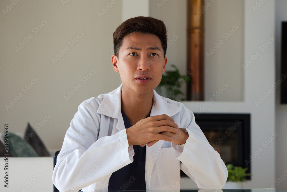 Obraz premium Mid-adult Asian man sitting at glass-topped table in modern living room wearing white lab coat