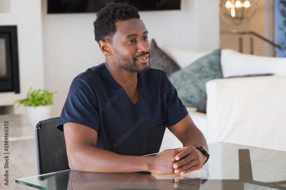 Obraz premium African American man in 30s sitting at glass table in living room, wearing navy scrubs, smartwatch