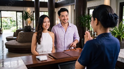 Smiling asian couple checking in at a luxury hotel reception desk. Cheerful tourists greeted by a hospitable receptionist during their summer holiday vacation