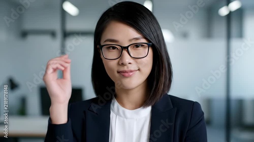A smiling woman with black hair wears glasses and a blazer. She holds up her hand near her face