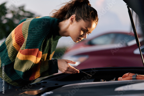 Quadro su tela A focused woman inspects a car engine under the open hood, showing concern and curiosity during roadside maintenance