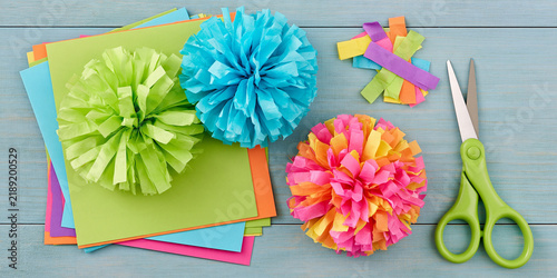 Crafting colorful tissue paper pom-poms with scissors and colored paper. Three vibrant pom-poms in shades of green, blue.