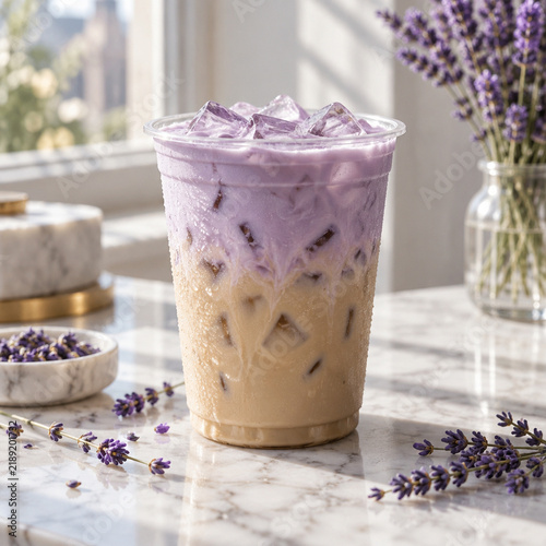 Iced lavender latte in a clear plastic cup with purple foam, served on a marble surface with fresh lavender sprigs and a vase in the background