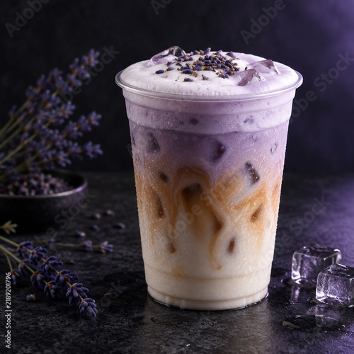 Iced lavender latte in a clear plastic cup with milk, coffee, and purple foam, garnished with dried lavender buds on a dark textured background